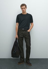 Schwarzes Rundhals-T-Shirt, dunkle Jeanshose, schwarzer Gürtel, trägt eine schwarze Stofftasche. Die Schuhe haben eine Wildleder-Textur mit einer sandfarbenen Gummisohle.