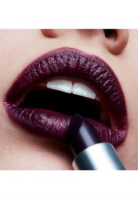Rouge à lèvres violet foncé appliqué sur des lèvres pleines, mettant en avant un fini brillant et des détails texturés. Des dents blanches sont visibles dans la bouche ouverte.