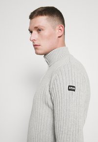 Jasnoszary sweter z prążkowanego materiału, z wysokim kołnierzem i naszywką z logo z lewej strony ramienia. Tekstura jest miękka i fakturowana.
