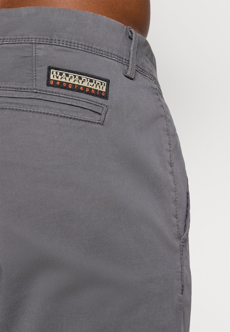 Graue Hose mit einer Gesäßtasche und einem rechteckigen, aufgenähten Stofflabel "Napapijri geographic" über der Tasche in der Nähe des Hosenbunds.