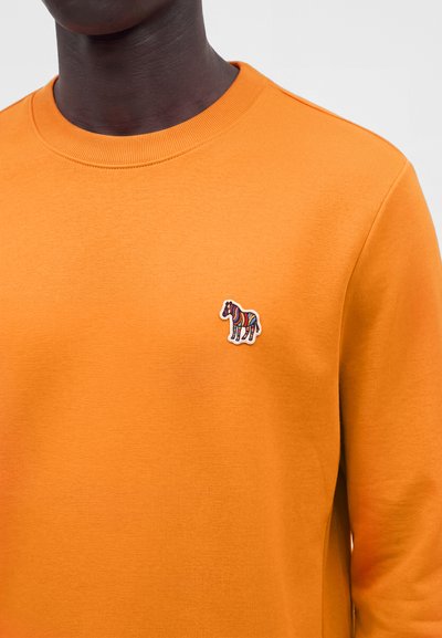 Personne à la peau foncée portant un t-shirt orange à col rond avec un petit patch zèbre coloré sur la poitrine gauche.