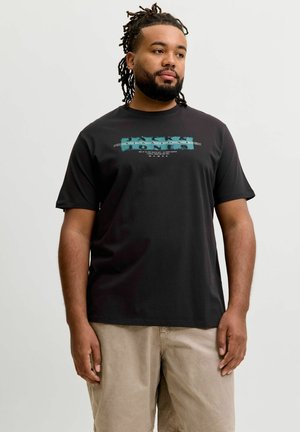 Mand med dreadlocks iført en sort grafisk T-shirt og beige bukser, stående mod en enkel lyse baggrund og kigger til siden.