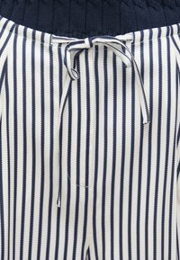Hlače na pruge tamnoplave i bijele boje s rebrastim tamnoplavim pojasom, opremljene vezicama za stezanje i glatkom teksturom.