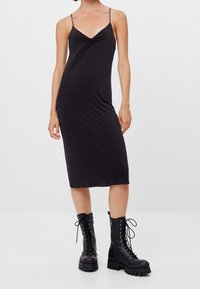 Robe midi noire à fines bretelles, avec un corsage froncé et une texture lisse ; associée à des bottes de combat noires à lacets avec une semelle épaisse.