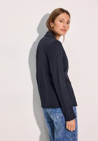 Marinblå jersey blazer med mjuk textur, enkel knapplåsning och snedskurna slag, ihopparad med ljusblå denimjeans.