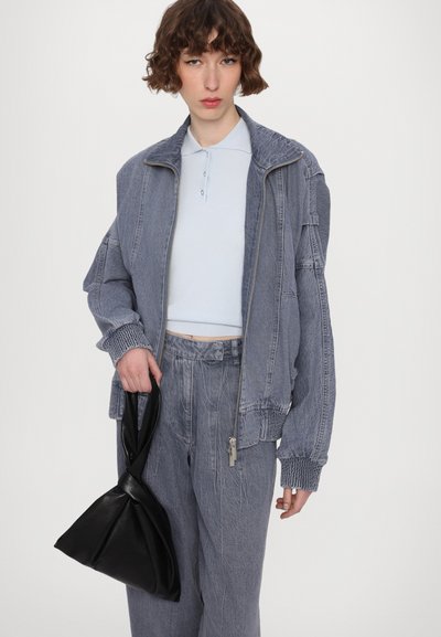 Jachetă din denim texturat gri, cu fermoar, manșete ribbate și pantaloni asortati. O geantă din piele neagră este ținută în mâna stângă.