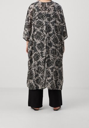Person iført en løs, lang kimono-stil top med blomsterprint i sort og grå over sorte vidde bukser og sandaler, stående på en ensfarvet baggrund.