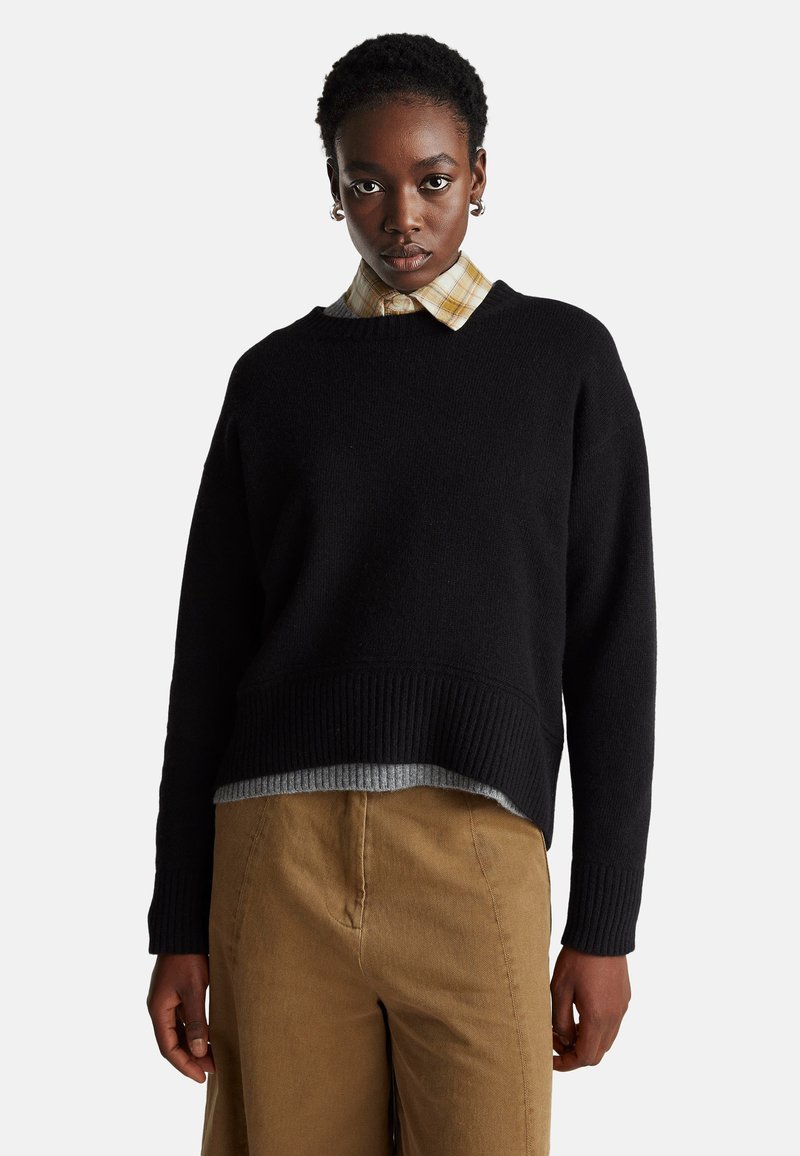 Schwarzer Strickpullover mit geripptem Saum, über einem grauen Hemd mit einem karierten Hemd in Gelb mit Kragen darunter getragen. Kombiniert mit hellbraunen Hosen.