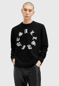 Sort sweater med hvid tekst i et cirkulært mønster, rund hals, lavet af et blødt, struktureret materiale. Sorte bukser er synlige nedenfor.