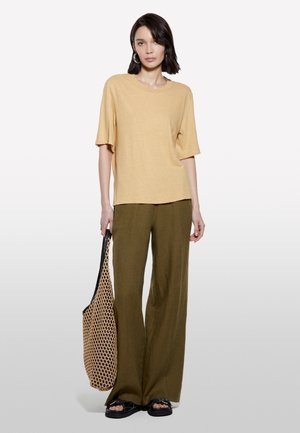 Top beige a maniche corte con una vestibilità comoda, abbinato a pantaloni a gamba larga verde oliva. La modella tiene una borsa in rete strutturata. Sandali neri.