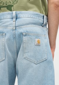 Φωτεινά μπλε τζιν από τζιν με πίσω τσέπες, με ένα μοτίβο λογότυπου Carhartt στην δεξιά τσέπη, φορεμένα με ένα πράσινο πουκάμισο και ορατό χέρι.