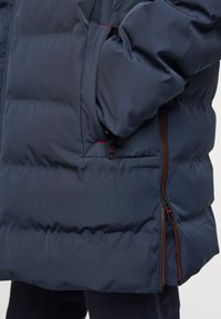 Giacca imbottita blu navy con superficie testurizzata, dotata di una tasca con zip e dettagli in zip arancioni a contrasto.