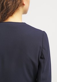 Námořnická modrá blůza s kulatým výstřihem a hladkým, lehkým materiálem, mírně nabranými rukávy a elegantním, přiléhavým střihem.