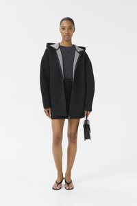 Zwart oversized hoodie met grijze accenten, ritssluiting aan de voorkant, textiel met structuur. Gecombineerd met een donkere top, zwarte rok en zwarte handtas. Sandalen zijn zichtbaar.