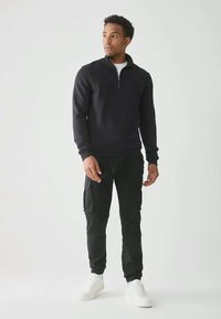 Muž stojící, oblečený v černém svetru s čtvrtinovým zipem, černých cargo kalhotách a bílých teniskách, na prostém světlém pozadí.