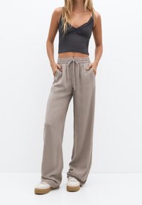 Světle šedé kalhoty s volným střihem, šňůrkou v pase a bočními kapsami, vyrobené z hladké, jemné tkaniny. Nosí se s tmavým crop topem s krajkovým lemem.