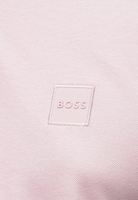 Rožinė tekstilė su subtilia tekstūra, centre pavaizduotas kvadrato formos įgaubtas logotipas „BOSS“. Audinys lygus ir švelniai atspindintis šviesą.