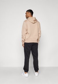 Beige Kapuzenpullover mit Kordelzugkapuze, kombiniert mit schwarzen Jogginghosen und weißen Turnschuhen. Glatte Textur und lässige Passform. Keine sichtbaren Muster.
