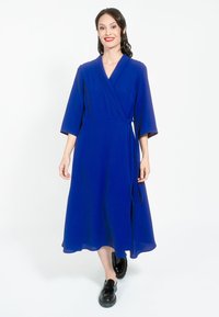 Plava haljina s omotom i rukavima do tri četvrtine, A-linija, završeni rub i pojas za vezanje. Nosena uz crne platforme.