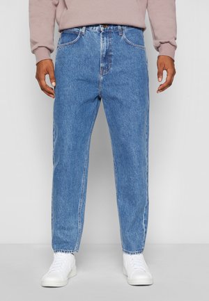 Personne portant un jean bleu clair à jambes droites, des baskets blanches et un sweat-shirt rose clair, debout devant un fond uni.