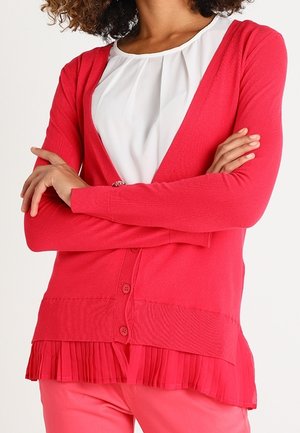Frau trägt eine rote, geknöpfte Strickjacke über einer weißen Bluse, die Arme verschränkt und korallrosa Hosen sichtbar.