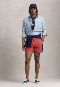 Cămașă din denim cu mânecă scurtă, polo în dungi alb-albastre, pantaloni scurți roșii, pulover bleumarin legat în jurul taliei și pantofi din pânză albastre.
