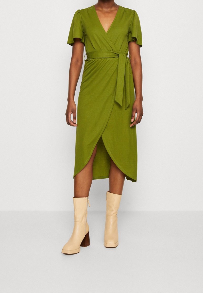 Personne portant une robe portefeuille verte à manches courtes et des bottes beige à talons mi-mollet, debout sur un fond clair uni.