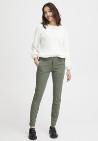 Krémový rebrovaný sveter s okrúhlym výstrihom v kombinácii so zelenohnedými priliehavými nohavicami. Obuv je čierna s lesklým povrchom.