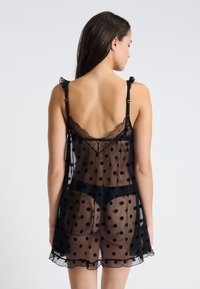 Kobieta widziana z tyłu, ubrana w czarną przezroczystą sukienkę w kropki, noszącą dopasowaną czarną bieliznę, na jednolitym jasnym tle.