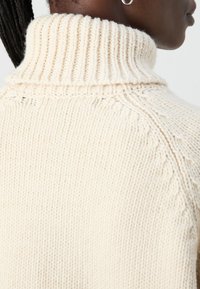 Cremefarvet strikket sweater med høj ribbet krave og teksturerede søm detaljer langs skuldrene og halsudskæringen.