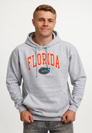 Ung mand iført en lysegrå hættetrøje med "Florida" og en grøn alligatorlogo, stående mod en ensfarvet hvid baggrund.