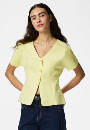 Femme aux longs cheveux foncés portant un chemisier jaune à manches courtes boutonné et un jean bleu foncé, debout, les mains derrière le dos.