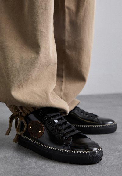 Schwarze Lackleder-Sneaker mit eingesticktem Rand, runder Zehenpartie und bracher Logopatch. Getragen mit beigen, umgeschlagenen Hosen, die eine lockere Passform zeigen.
