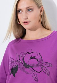 Paarse katoenen T-shirt met een zwart bloemenontwerp met glinsterende accenten, ronde halslijn en lange mouwen.