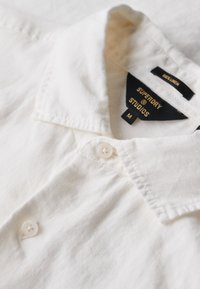 Camisa blanca de lino con cuello abotonado, que presenta un solo botón y una etiqueta que muestra "Superdry Studios" y las etiquetas de cuidado.