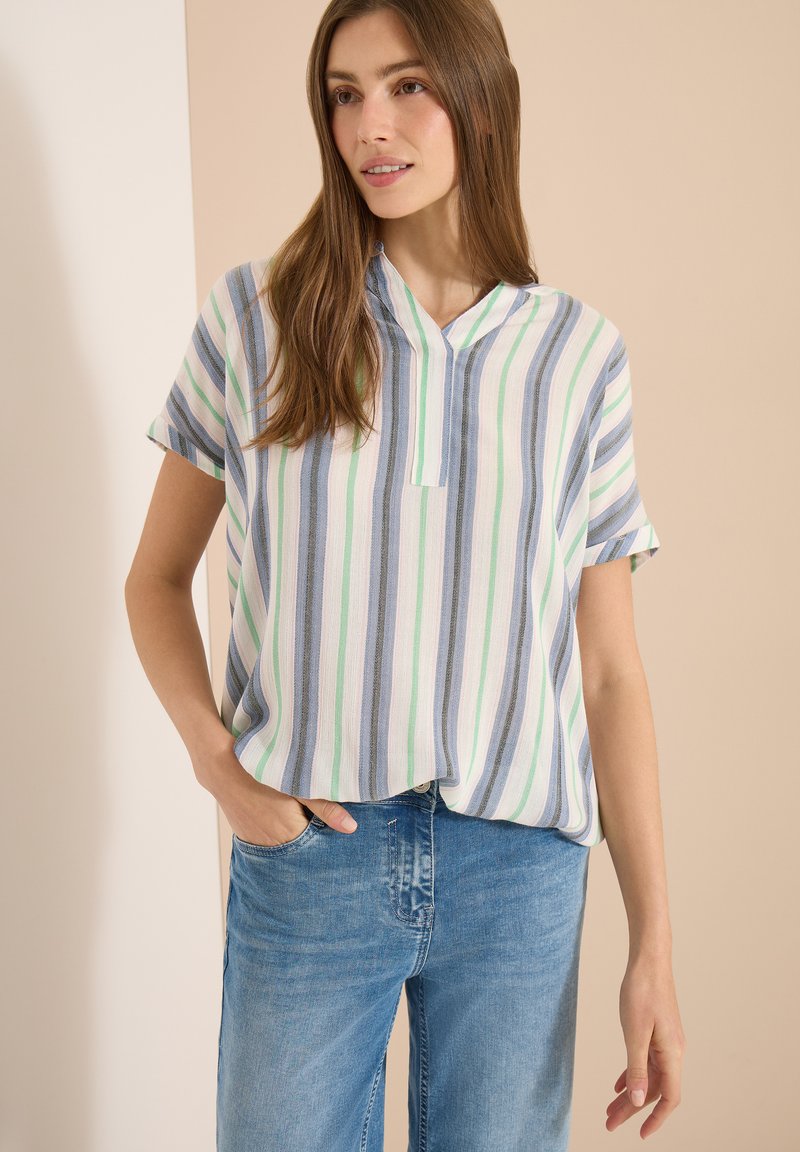 Jeune femme aux longs cheveux bruns portant un haut blanc à manches courtes avec des rayures verticales bleues et vertes et un jean bleu.