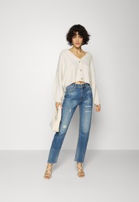 Suéter creme cropped com relevo e cinco botões, jeans reto de perna longa desfiados em azul médio, sapatos de salto brancos com tiras e uma bolsa branca.