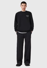 Young man wearing black AllSaints sweatshirt and loose black pants, standing with hands in pockets against light gray background.