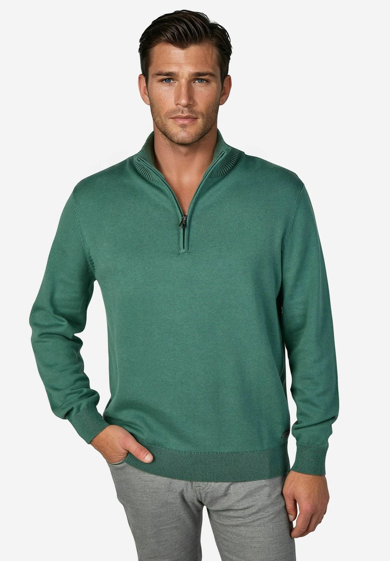 Grüner Strickpullover mit einem Viertelreißverschluss am Kragen, gerippten Bündchen und Saum. Glatte Textur mit figurbetonter Silhouette, getragen über hellgrauen Hosen.