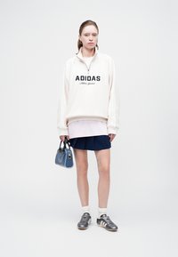 Felpa bianca con zip e logo nero "ADIDAS Athletic Garment", indossata sopra una maglietta rosa, abbinata a una gonna plissettata blu navy e sneakers argentate.