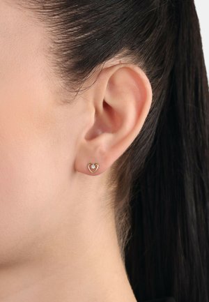 Pendientes en forma de corazón de oro con un centro de gema clara, que presentan un acabado suave y un diseño abierto, exhibidos en la oreja de un modelo.