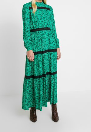 Robe maxi à motifs verts avec des manches longues, accents en dentelle noire et design en volants. Associée à des bottines à texture marron.