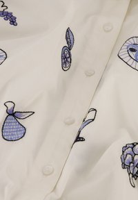 Witte blouse met meerkleurige geborduurde ontwerpen van fruit en bloemen in tinten blauw en paars, met doorzichtige kunststof knopen.