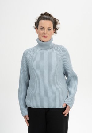 Vrouw met krullend donker haar, gekleed in een lichtblauwe geribbelde coltrui en zwarte broek, staand tegen een effen witte achtergrond.