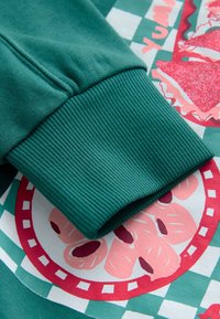 Zelený manžet mikiny so žebrovanou textúrou, prekrytý farebnou grafickou potlačou s motívmi jedla a károvaným vzorom.