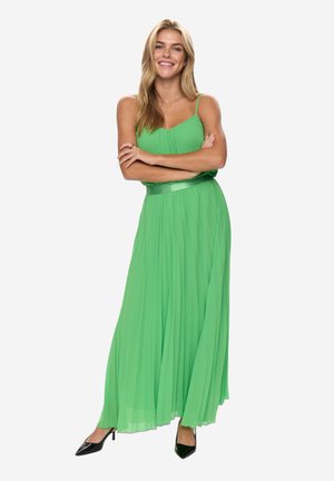 Smilende kvinde med langt blondt hår, iført en ærmeløs grøn maxi-kjole og sorte hæle, stående med armene over kors mod en hvid baggrund.