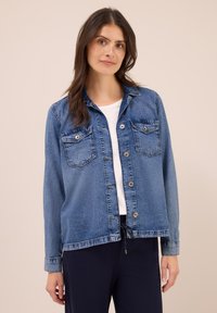 Vrouw met lang bruin haar die een knoopblauw denimjack over een witte blouse en donkere broek draagt, tegen een beige achtergrond.