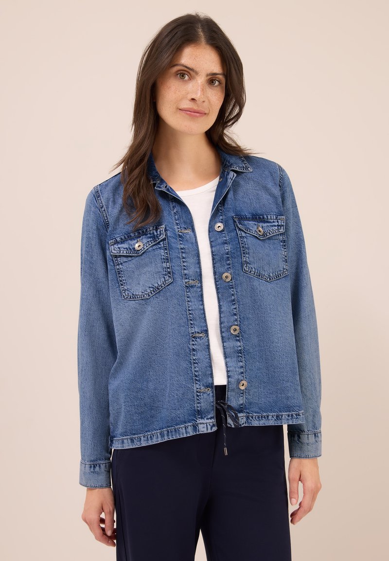 Donna con lunghi capelli castani che indossa una giacca di jeans blu con bottoni sopra una camicia bianca e pantaloni scuri, su uno sfondo beige.