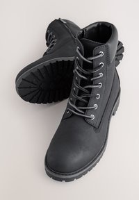 Schwarze Lederknöchelstiefel mit robusten Gummisohlen, rundem Zeh und grauen Schnürsenkeln. Mit einem seitlichen Reißverschluss für einfaches An- und Ausziehen.