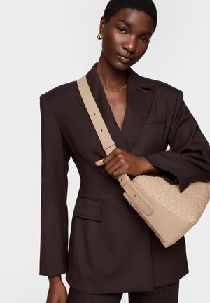 Kobieta w ciemnobrązowej marynarce i złotych kolczykach kołach, trzyma beżową torebkę o fakturowanej powierzchni z regulowanym paskiem przewieszonym przez pierś.